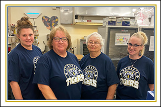 4 ladies in blue t-shirts smiling behind a lunch prep table